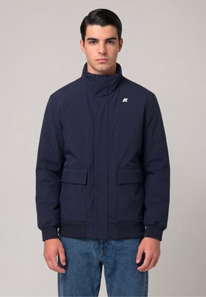 Jonge man met donker glad naar achteren gekamd haar, draagt een marineblauw jasje met voorzakken en een blauwe spijkerbroek, staand tegen een effen lichte achtergrond.