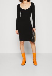 Robe noire à manches longues avec un décolleté en V blanc, design ajusté et texture lisse. Associée à des bottines orange et portant un sac.