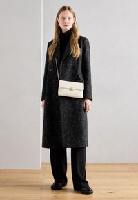 Manteau long et surdimensionné à motifs noirs et blancs, associé à un sac bandoulière crème avec une boucle circulaire. Pantalon et chaussures foncés.