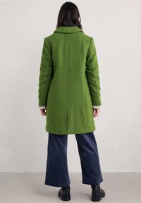 Zielony, teksturowany płaszcz z wyraźnym kołnierzem, pokazany od tyłu. Połączony z ciemnymi, szerokimi spodniami z korduroyu i czarnymi butami.