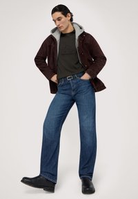 Mörkbrun mockajacka över en grå hoodie, grön skjorta med tryckt mönster, blå vida jeans och svarta stövlar.