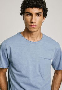 Šviesiai mėlynas trumpomis rankovėmis marškinėliai su apvalia kaklo iškirptele, pasižymintys krūtinės kišene ir subtiliu tekstūriniu raštu visame audinyje.