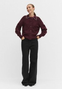 Bordowy kardigan dzianinowy z przodem zapinanym na guziki i kołnierzem ozdobionym małymi białymi akcentami, w połączeniu z czarnymi spodniami z szerokimi nogawkami.