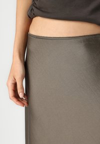 Knee-pikkuses oliiviroheline seelik, mis on valmistatud siledast ja kergest kangast, kitseneva vöökoha ning peene läikega, käsi seeliku külje peal.
