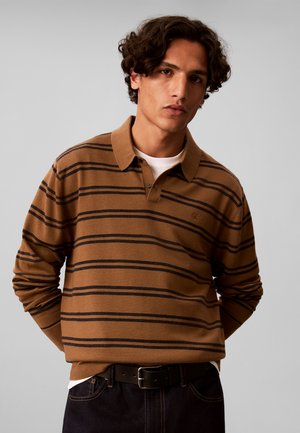 Junger Mann mit lockigem dunklem Haar, der ein braun-schwarz gestreiftes langärmeliges Poloshirt und dunkle Hosen trägt und mit den Händen hinter dem Rücken steht.