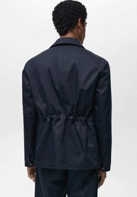 Giacca leggera blu navy con vita regolabile, texture morbida e polsini con bottoni; caratterizzata da un colletto classico e dettagli minimi.