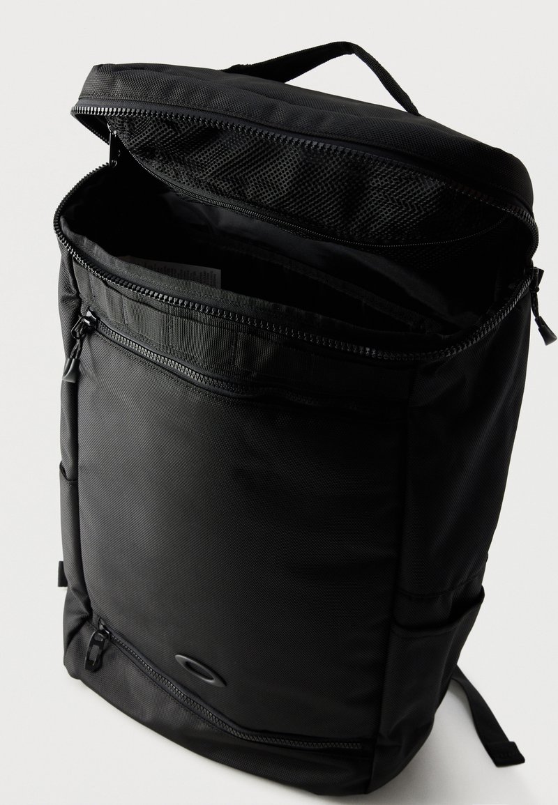 Sac à dos noir avec compartiment principal ouvert, poche zippée à l’avant, poignée supérieure et bretelles rembourrées sur fond blanc.