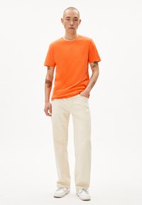 T-shirt en coton orange avec un col rond, associé à un pantalon droit de couleur crème. Des baskets blanches complètent la tenue.