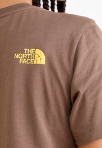 Hnědé bavlněné tričko s krátkým rukávem, na zadní straně s žlutým logem The North Face, hladká textura.