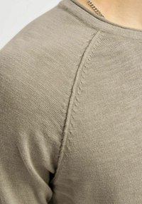Maglione a maglia color taupe chiaro con una texture liscia, caratterizzato da maniche raglan e cuciture dettagliate lungo le cuciture.