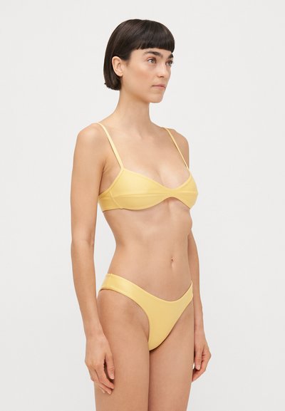 Set bikini giallo caratterizzato da un design minimalista con spalline sottili, un bralette triangolare e un pantaloncino a vita bassa coordinato, realizzati in una texture liscia.