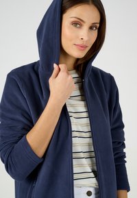 Sudadera de forro polar azul marino con cremallera en la parte delantera y capucha forrada, llevada sobre una camiseta de rayas. La textura parece suave y cálida.