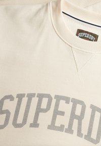Sweat-shirt écru avec col rond côtelé, impression du texte "SUPERDRY" en gris, et tissu texturé avec un léger éclat.