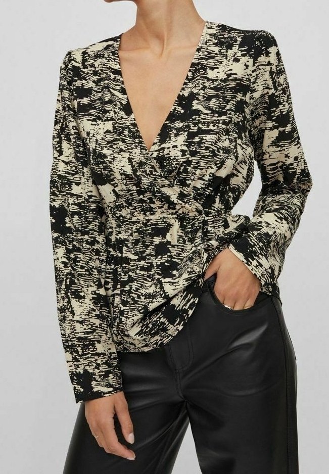 Femme portant un chemisier portefeuille à manches longues avec un imprimé abstrait noir et beige, associé à un pantalon en cuir noir, main dans la poche.