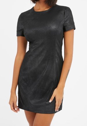 Femme portant une mini-robe noire moulante à manches courtes avec un tissu texturé, debout devant un fond blanc uni.