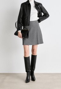 Donna che indossa una giacca di pelle nera, un dolcevita bianco a costine, una gonna a scacchi bianco e nero, stivali neri fino al ginocchio e tiene una borsa nera.