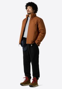 Braune isolierte Jacke, grauer Sweatshirt, schwarze Hose und braune Stiefel mit roten Schnürsenkeln. Einfaches Design mit funktionalen Details.