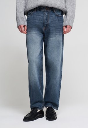 Persona che indossa jeans blu a vestibilità ampia, maglione grigio lavorato a maglia, mocassini in pelle nera e calzini bianchi, in piedi davanti a uno sfondo grigio uniforme.