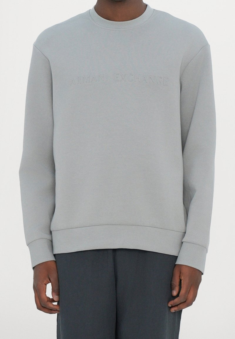 Homme portant un sweat-shirt gris clair avec un logo en relief "Armani Exchange" sur la poitrine et un pantalon foncé sur fond blanc.