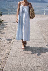 Vaaleansininen, ilman hihoja oleva maksimekko rennolla istuvuudella, yhdistettynä valkoisiin flip-floppeihin ja kudottuun olkipussiin. Kivetyllä polulla järven vieressä.