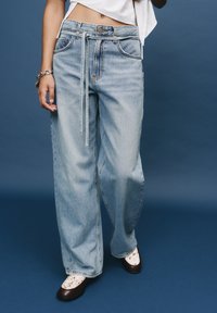 Hellblaue weite Jeans mit gebundenem Bund, ausgestattet mit Vordertaschen und leichtem Verblassen. Getragen mit gemusterten Schuhen. Der Hintergrund ist blau.