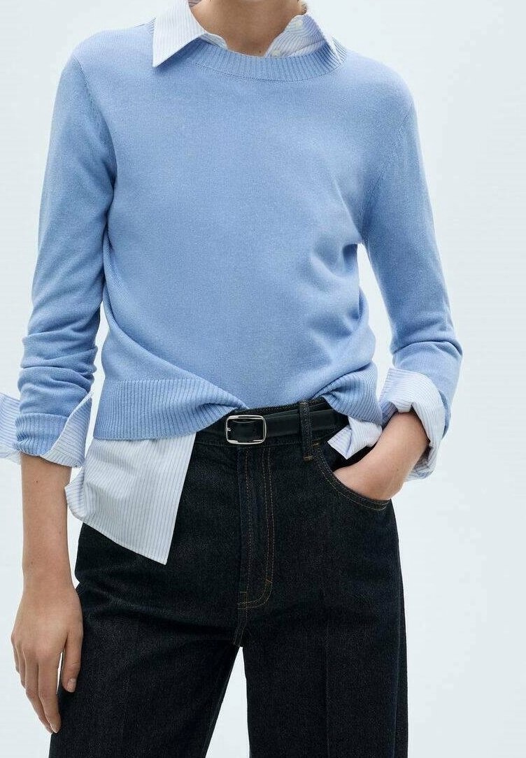 Personne portant un pull bleu clair par-dessus une chemise blanche à col avec les manches retroussées, rentrée dans un pantalon noir avec une ceinture noire, main dans la poche.