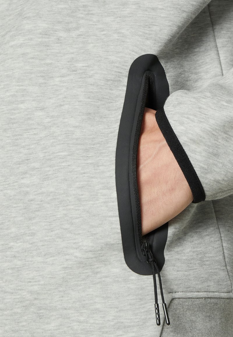 Tejido de sudadera gris con un bolsillo con cremallera. La cremallera y el contorno del bolsillo son negros, lo que realza la funcionalidad. Se puede ver la textura suave.