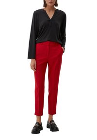 Zwarte blouse met lange mouwen en V-hals, gecombineerd met felrode, op maat gemaakte broek. Model draagt zwarte instappers met metalen accenten.