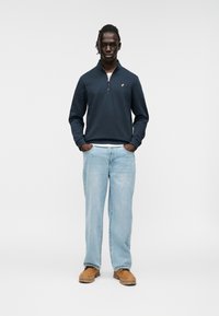 Ναυτικό φούτερ με φερμουάρ και ψηλό γιακά, μπεζ ραφές, σε συνδυασμό με ανοιχτό μπλε τζιν και καφέ παπούτσια, με μινιμαλιστικό σχεδιασμό.