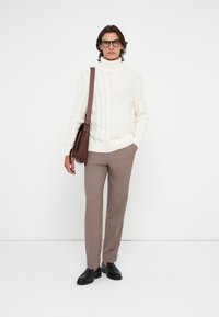 Mann in weißem, kabelgestricktem Rollkragenpullover, beigen Hosen, schwarzen Slippern, mit Brille und einer braunen Lederumhängetasche.