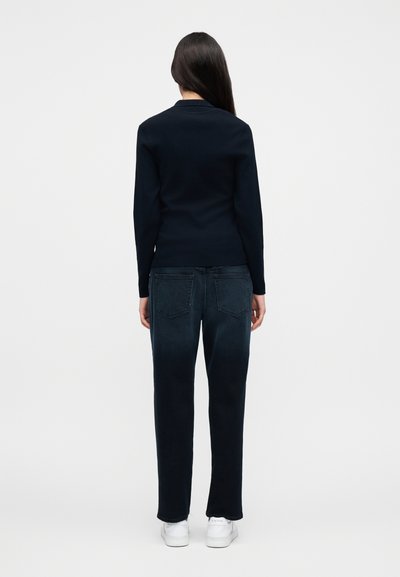 Γυναίκα που στέκεται με την πλάτη γυρισμένη, φορώντας μαύρο μπλουζάκι με μακριά μανίκια, σκούρο μπλε τζιν και λευκά αθλητικά παπούτσια σε απλό φωτεινό φόντο.