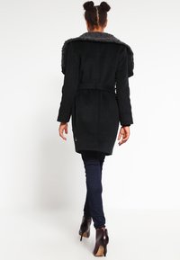 Cappotto nero con colletto in pelliccia, vita con cintura e maniche lunghe. Indossato sopra jeans scuri e stivali con tacco alto, che mettono in mostra un design elegante.