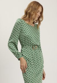 Zielona sukienka w wzory, wykonana z gładkiego materiału, z długimi rękawami, dekoltem w łódkę i paskiem w talii zakończonym okrągłą klamrą.