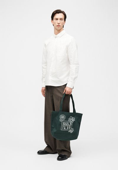 Mann som har på seg hvit skjorte med knapper og brune vidbukser, som holder en mørkegrønn totebag med hvit Maison Kitsuné-logo.