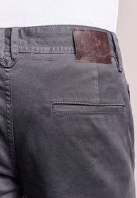 Grå bomullbyxor med en brun läderlappar på baksidan av midjan, med en enkel slitsficka, slät textur och avslutande sömmar.