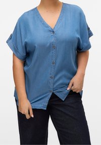 Person wearing a blue short-sleeve button-up shirt with rolled cuffs and dark pants, holding the shirt's hem on a white background.