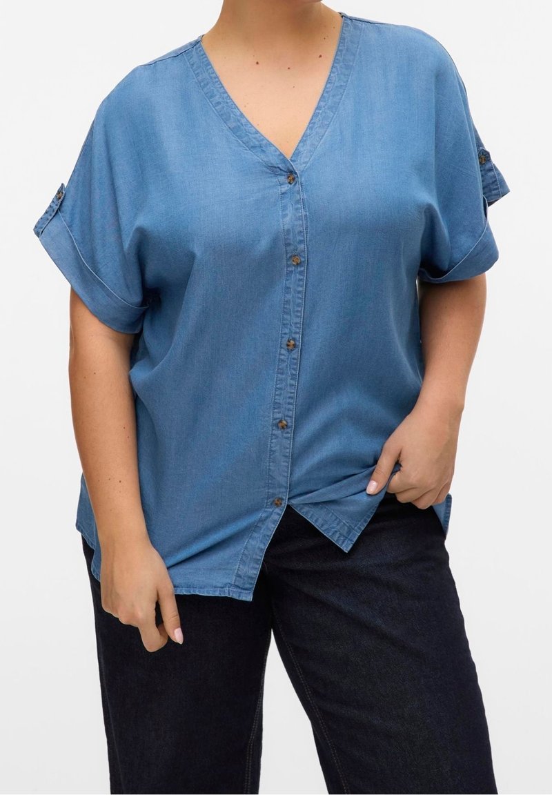 Person wearing a blue short-sleeve button-up shirt with rolled cuffs and dark pants, holding the shirt's hem on a white background.