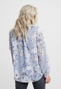 Blusa floreale blu, realizzata in un tessuto leggero e trasparente. Il design include grandi stampe floreali e maniche lunghe con polsini elastici.