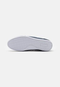 Semelle de sneaker Lacoste, présentant un dessous en caoutchouc blanc texturé avec un motif circulaire et un bord supérieur bleu foncé. Design simple et fonctionnel.