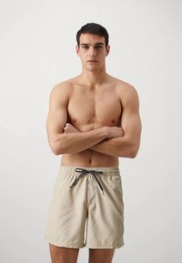 Ljusa beiga badshorts med elastisk midja, justerbar dragsko och sidofickor. Slät textur och knälång design.