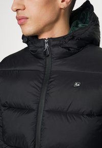 Giovane con giacca imbottita nera con cappuccio, zip frontale, piccolo logo sul petto e un orecchino a cerchio dorato nell’orecchio sinistro.