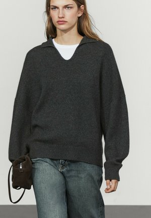 Jonge vrouw die een donkergrijze oversized trui draagt over een wit overhemd, lichtblauwe spijkerbroek en een kleine donkerbruine tas bij zich heeft.