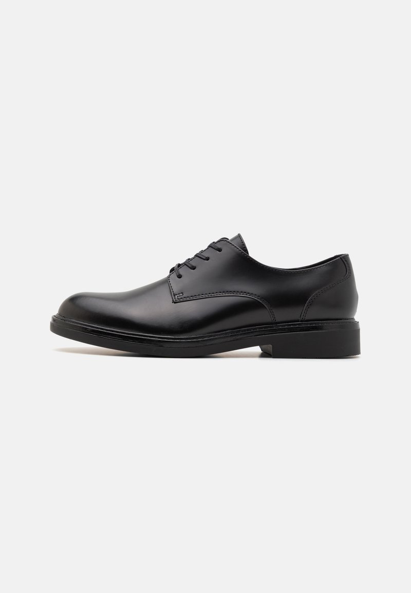 Chaussure habillée en cuir noir avec une finition lisse, bout rond, six œillets et un petit talon. Présente des coutures délicates et une semelle plate.