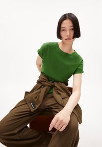 Camiseta de algodón verde con mangas cortas y cuello redondo, combinada con pantalones marrones de corte holgado atados a la cintura.