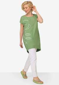 Zelená krátká tunika s bílým nápisem, v kombinaci s bílými kalhotami a zelenými klouzavými sandály. Hladký materiál a uvolněný střih.