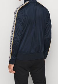 Marinblå atletisk jacka tillverkad av mjukt tyg med hög krage, randiga sidodetaljer med logotyper samt ribbade ärmslut och fåll.