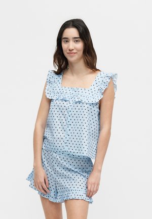 Νεαρή γυναίκα με ανοιχτό μπλε αμάνικο τοπ και σορτς με καφέ πουά και βολάν στα άκρα, στέκεται μπροστά σε απλό φόντο.