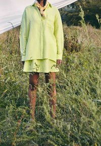 Chemise en lin vert lime à manches longues, avec fermeture par boutons et une poche, associée à une jupe superposée assortie. Portée avec une écharpe à motifs.
