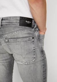 Grå denimjeans med en slät yta, med en svart läderlogotyp på baksidan av midjan och standard fickdesign.