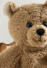 Bruine teddybeer van pluizige, gestructureerde stof. Heeft zwarte plastic ogen, een gestikte neus en een kleine bruine rugzakriem.